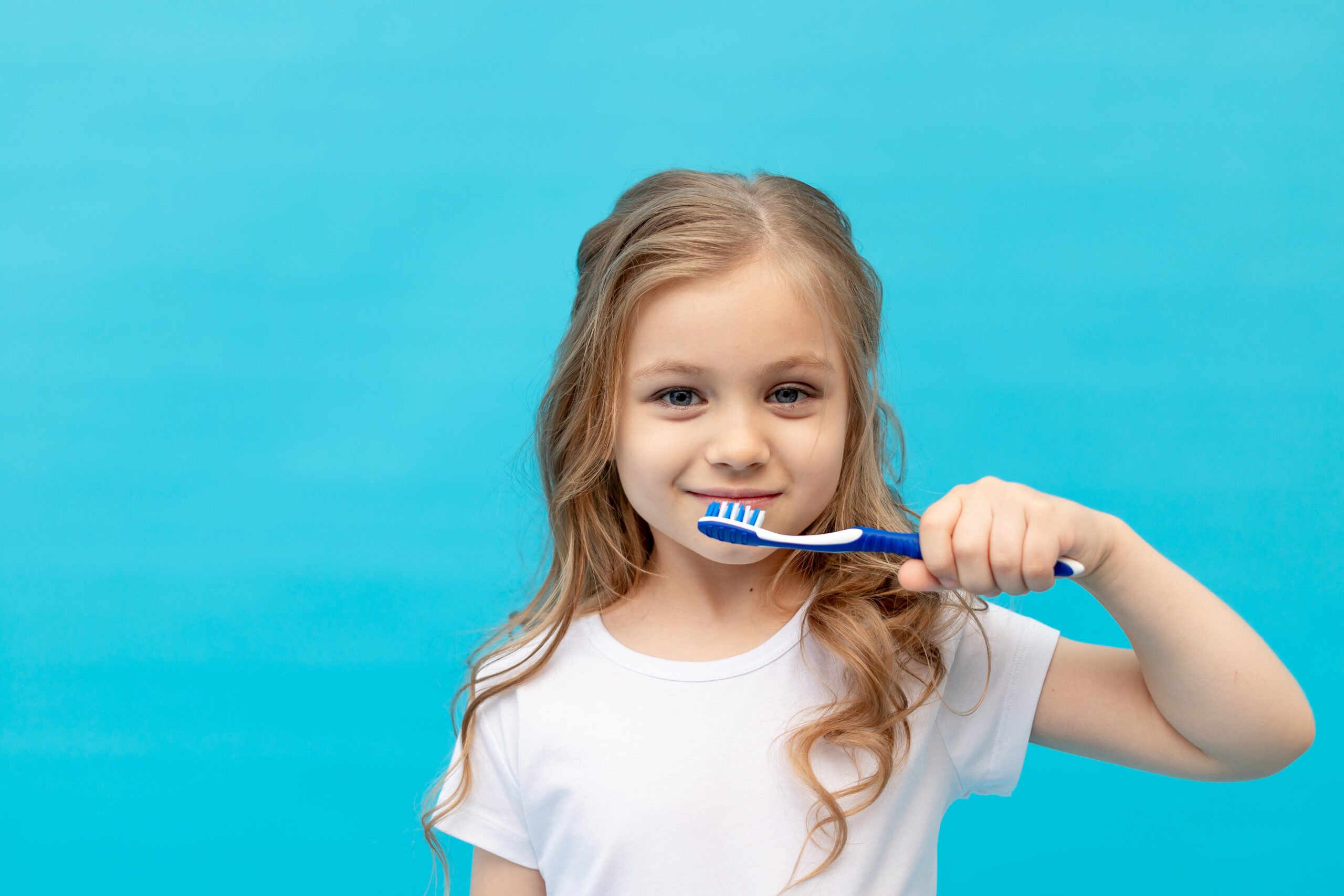 cute little girl child in a white T-shirt brushing her teeth with a toothbrush on a blue background, the concept of hygiene and dentistry, space for text